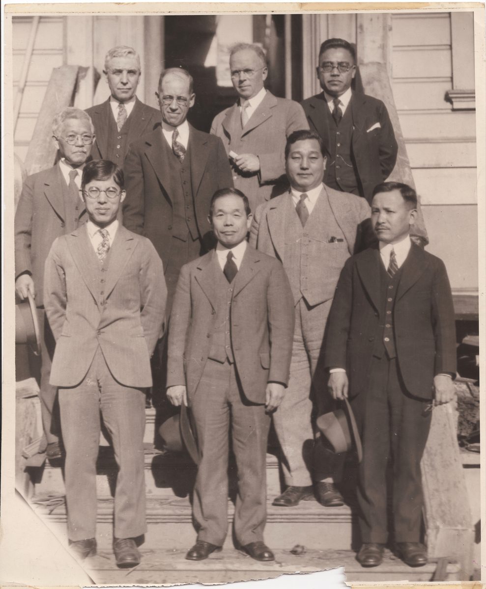Ten men in suits, American and Japanese, standing on the front steps of an old wooden building.