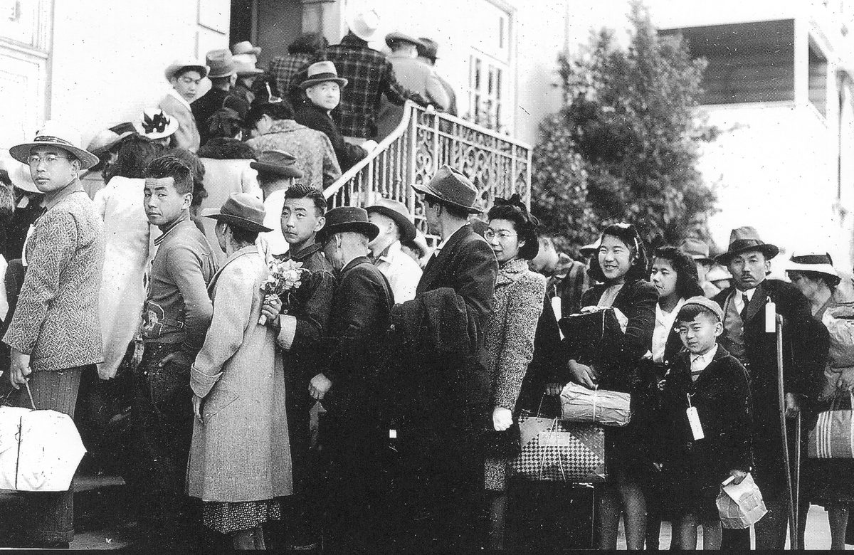 A crowd of Japanese American men, women and children in coats and hats, holding bags and bundles outside the YMCA.