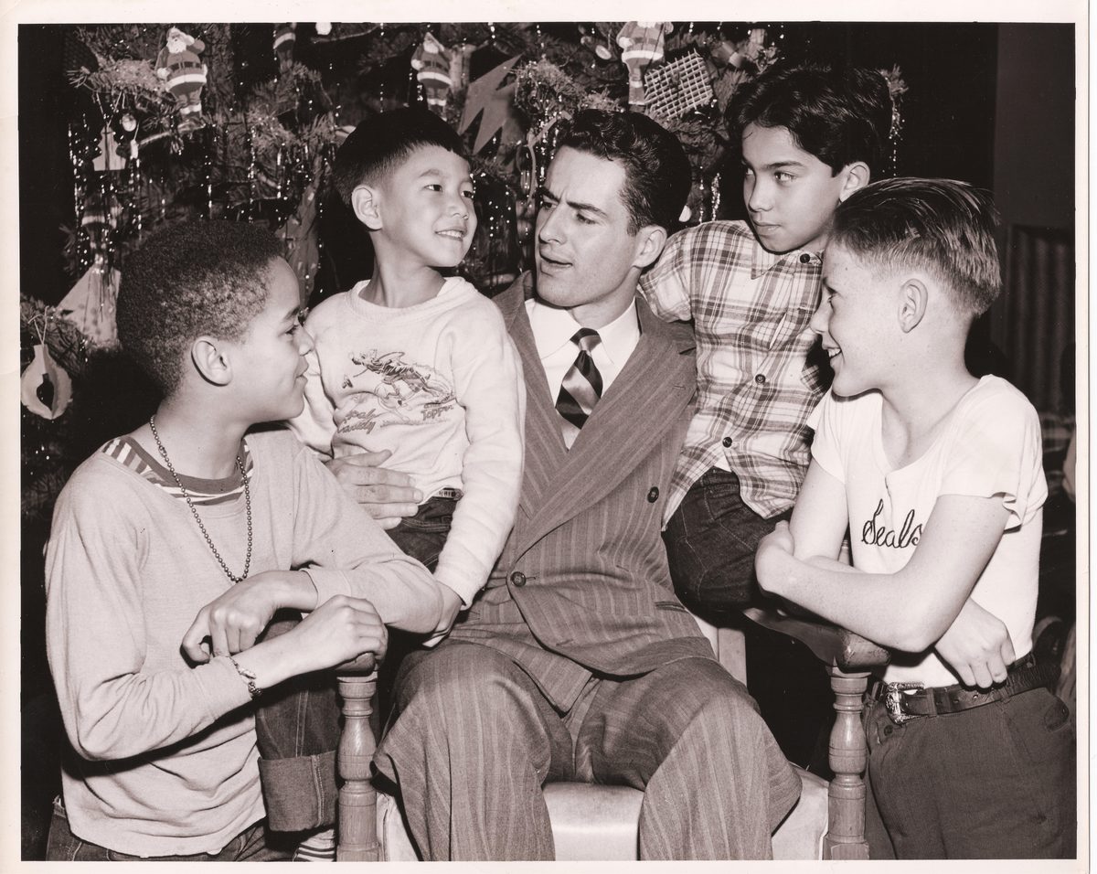 Four boys of different backgrounds laughing with a man in a pinstripe suit in front of a Christmas tree.