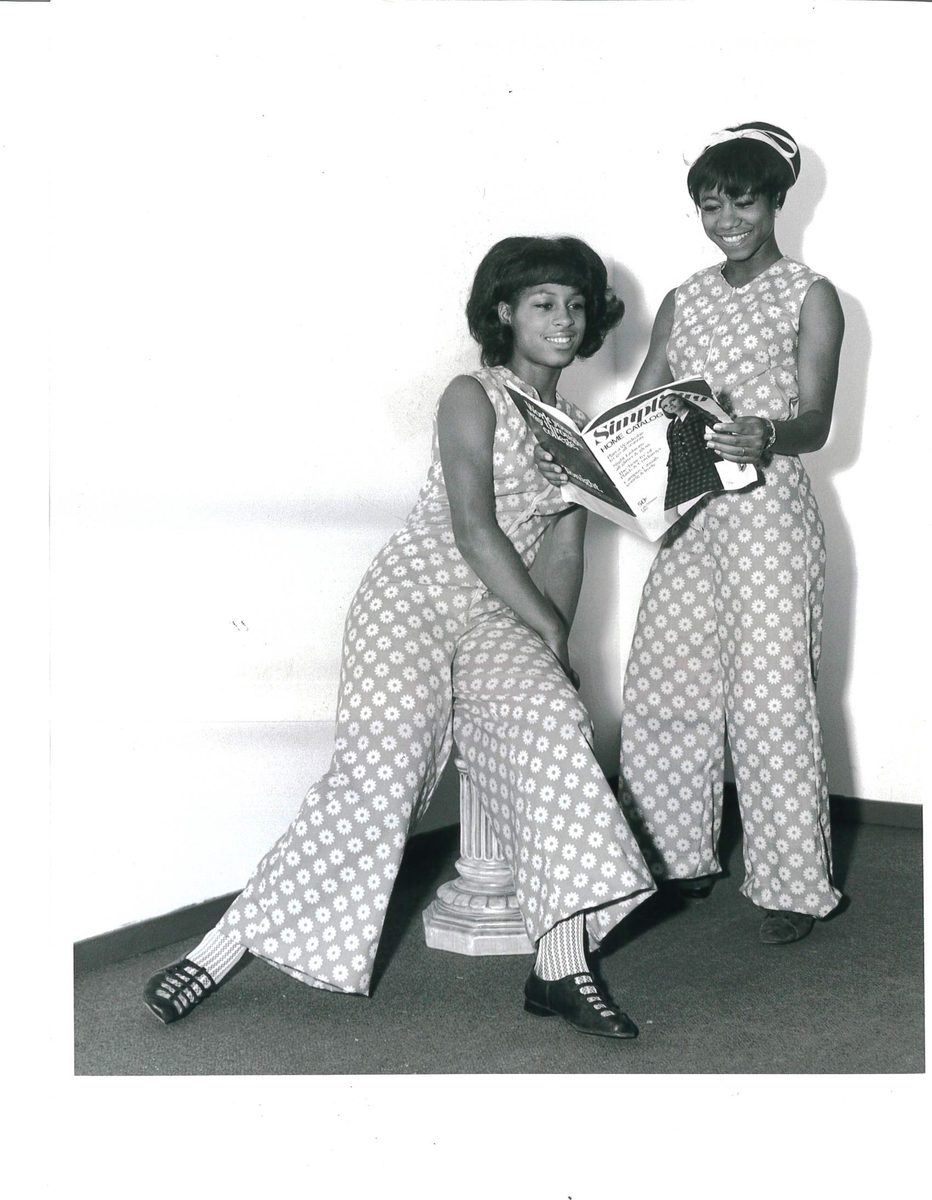 Two young women in matching patterned jumpsuits looking at a sewing-pattern magazine.